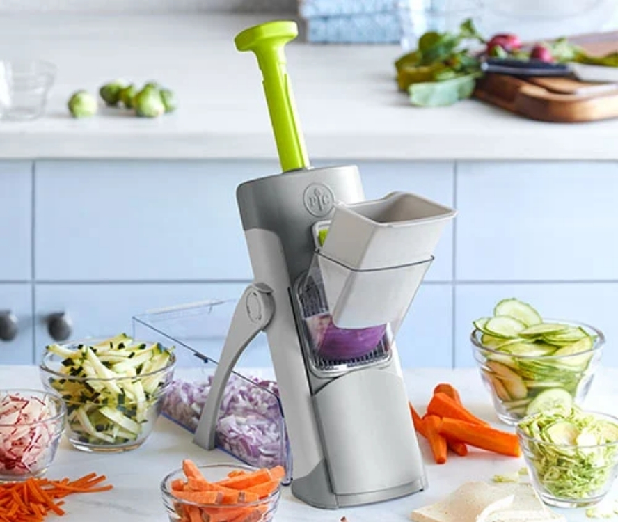 Image of Pampered Chef's Rapid-Prep Mandoline on a kitchen countertop, surrounded by thinly sliced vegetables in bowls.