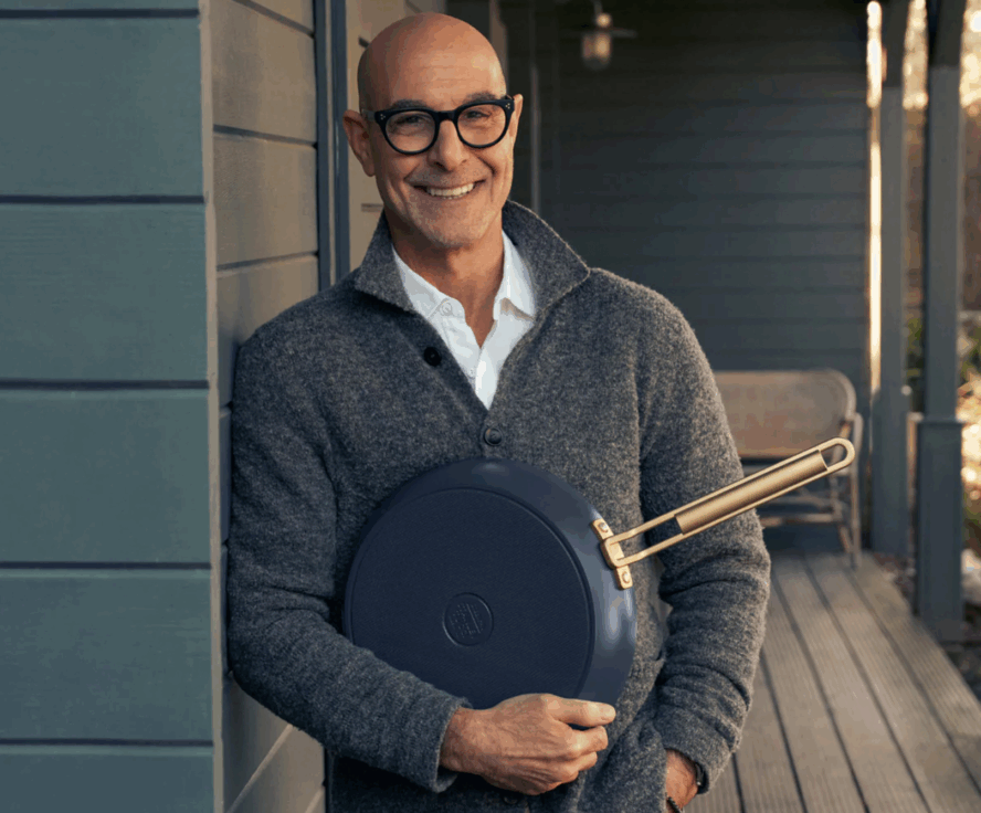 Image of Stanley Tucci holding a pan from his GreenPan collection and leaning against the exterior wall of a blue farmhouse.