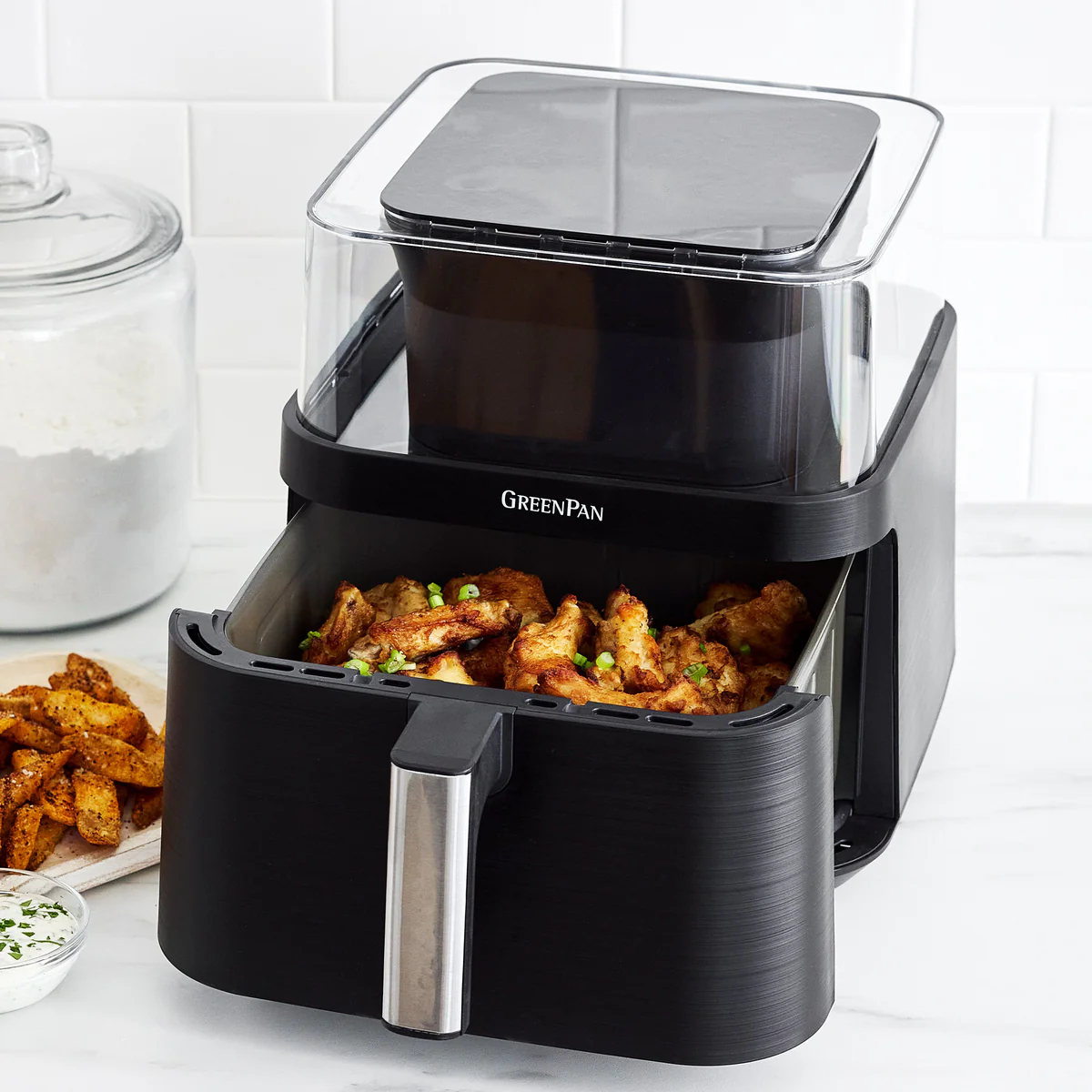 Image of the GreenPan Elite Air Fry Oven in Black, displaying a drawer full of chicken wings. It's set on a white marble countertop with various dishes surrounding it, including a pizza, croissants, stuffed peppers, a steak, salmon, and more.