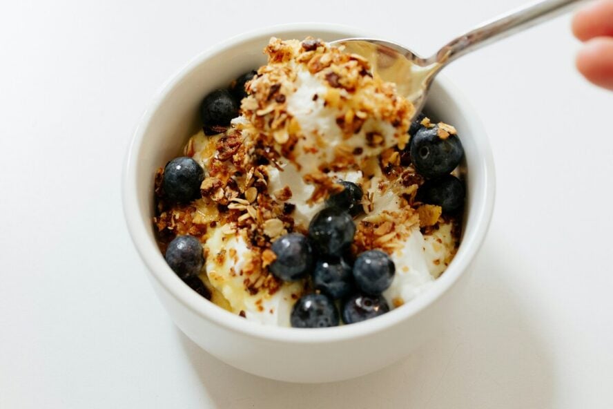 Image of a small bowl of Greek yogurt topped with granola and blueberries, with a spoon digging into the bowl.