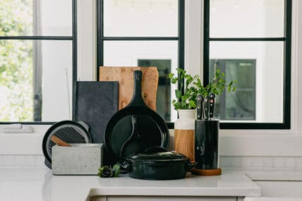 Image of black Xtrema pure ceramic cookware leaning against a kitchen wall of windows on a kitchen counter. The Xtrema cookware is srcd with other items like a wood cutting board and a natural vase filled with herbs.