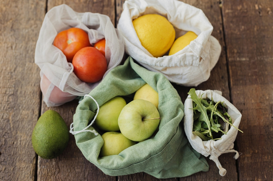 Image of several reusable grocery bags on a wooden table, filled with various produce items (tomatoes, lemons, arugula).