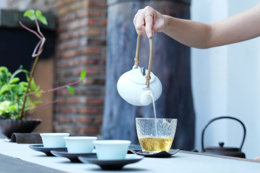 Image of a hand pouring tea from a kettle into a glass mug, in front of several other ceramic cups. Brewing loose-leaf tea is a great way to avoid a tea bag's microplastic content.