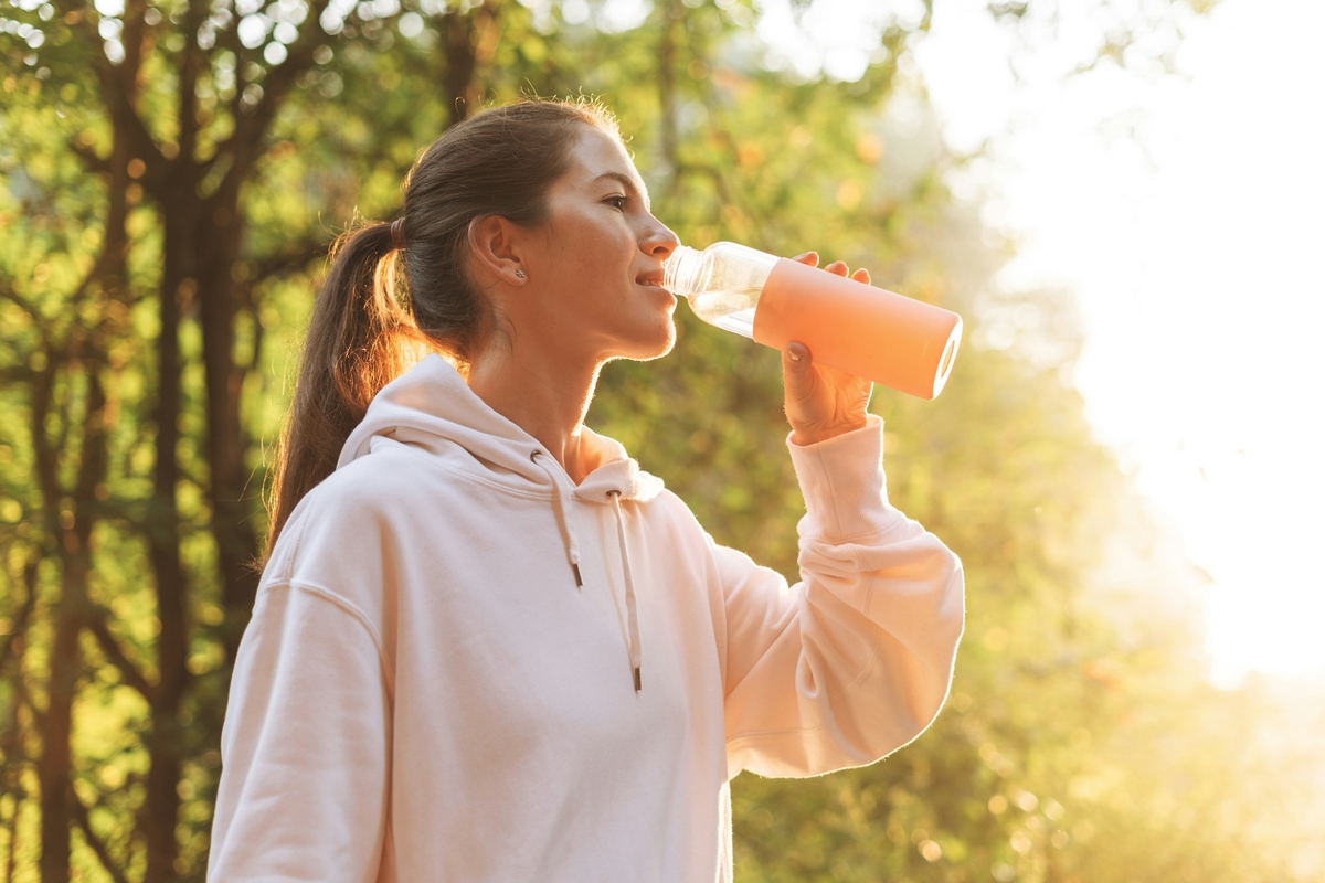 Image of a young brunette woman in a sweatshirt and ponytail, drinking water from a sports water bottle.