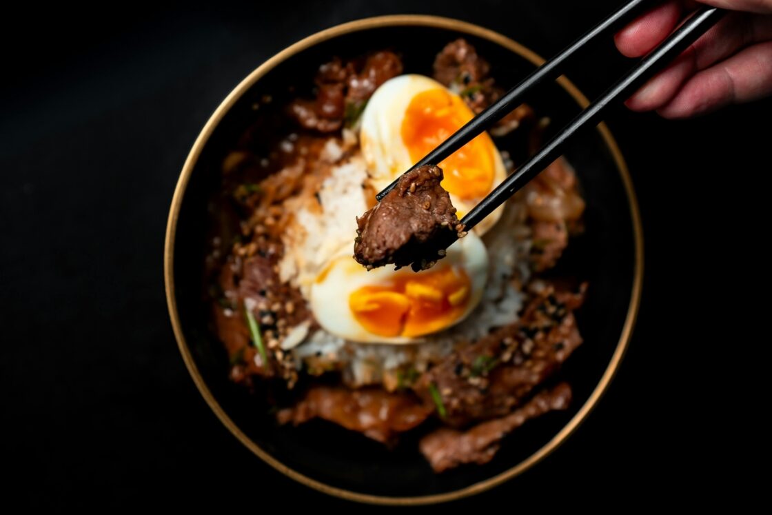Image of a pair of chopsticks with a bite of beef hovering over an Asian beef bowl with eggs and rice.