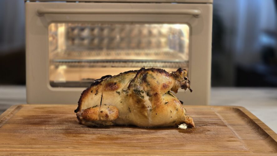 Image of a whole roasted chicken on a cutting board in front of a air fryer 6-1 Wonder Oven by Our Place