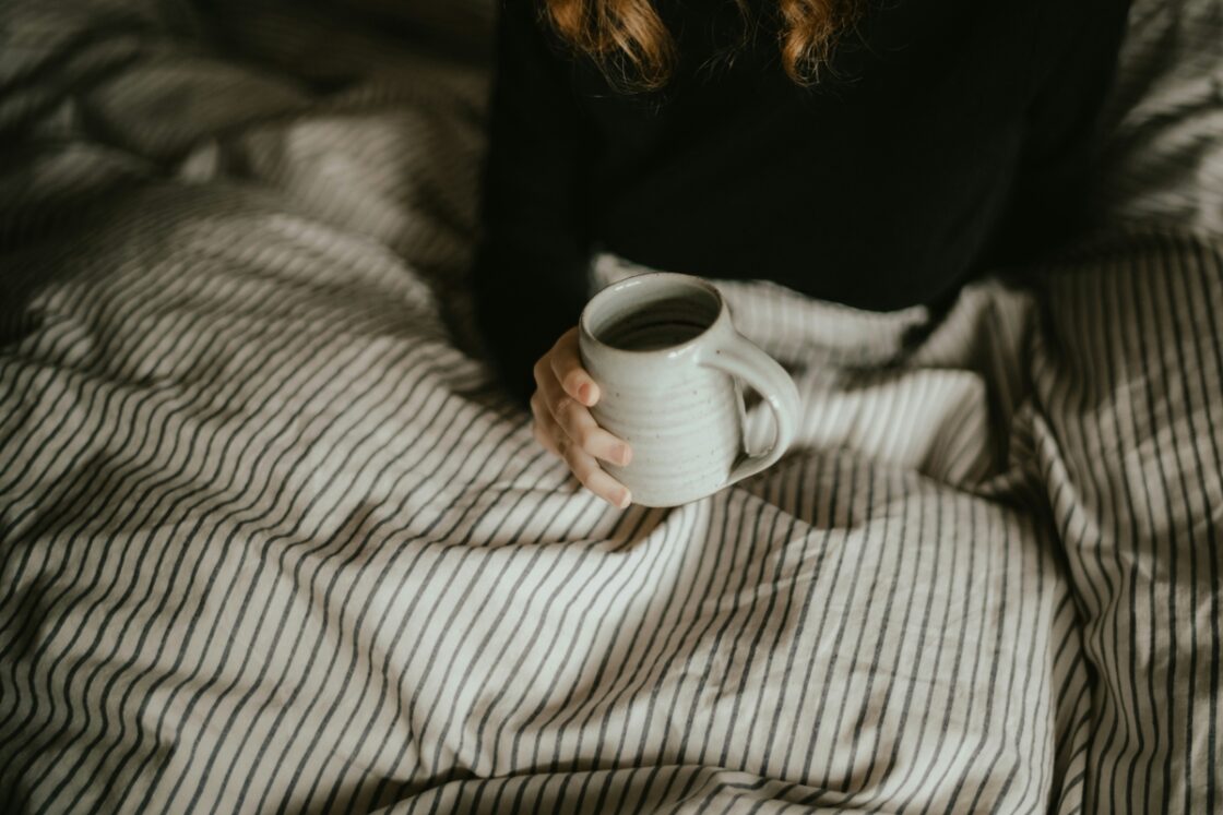 Image of a person sitting in bed under cozy sheets, holding a mug of coffee or tea.