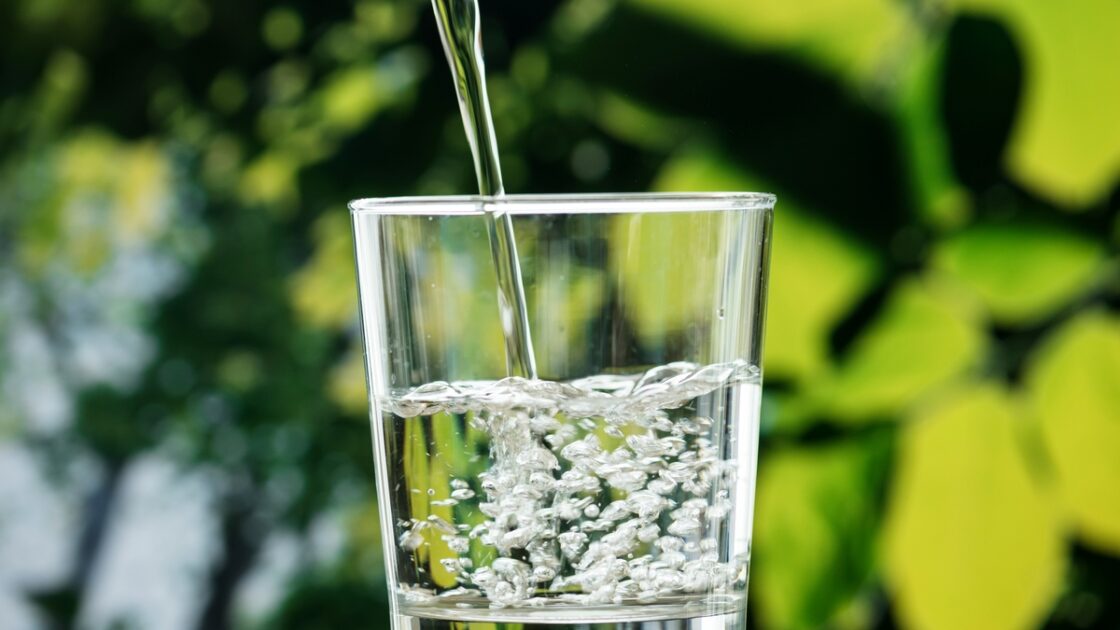 Image of water being poured into a glass set against a background of lush greenery.