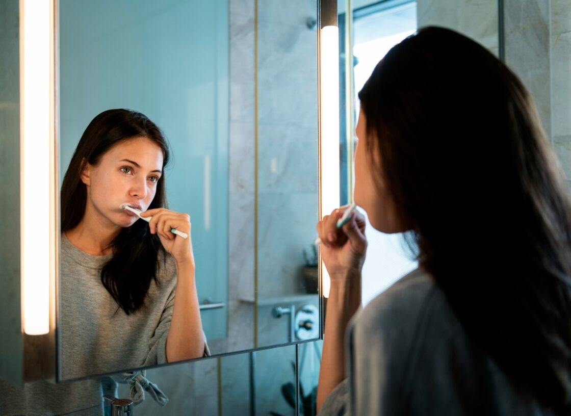 Image of a young woman brushing her teeth and looking at herself in the mirror.