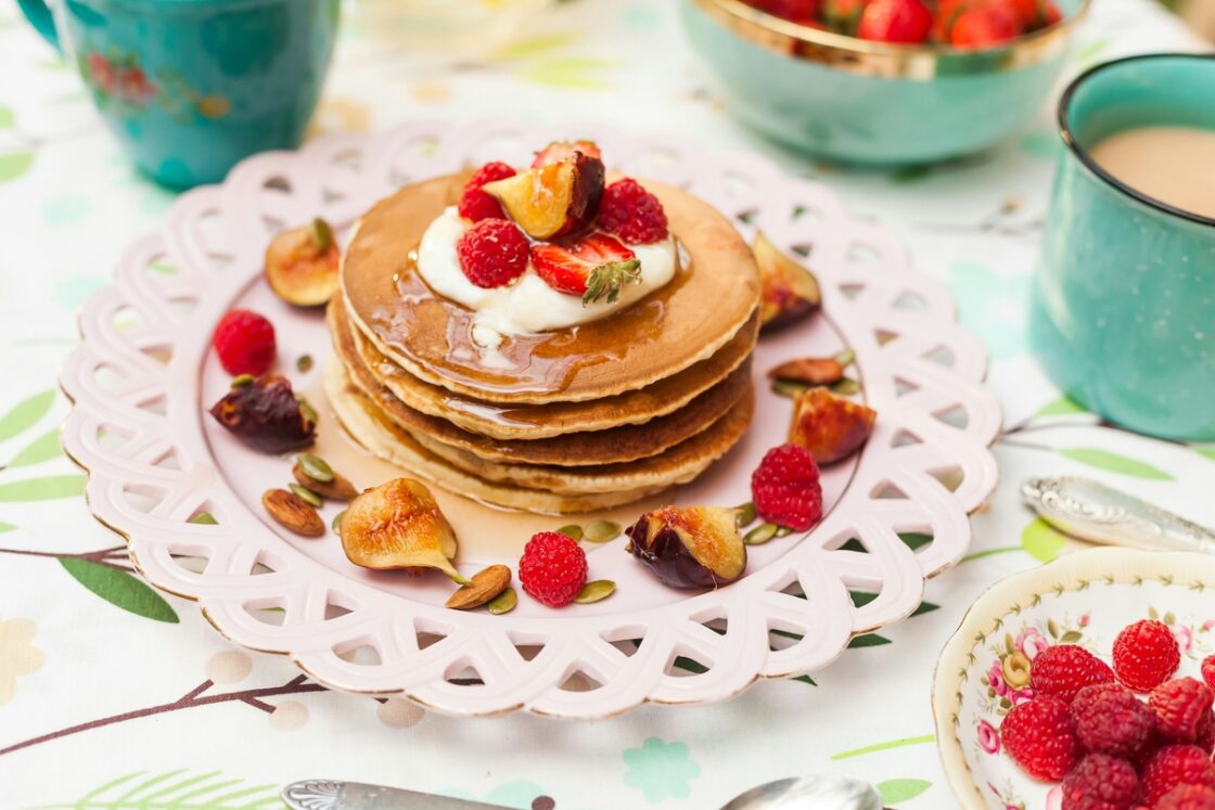 Image of a stack of pancakes topped with greek yogurt, raspberries, figs, and syrup on a cute pink plate.