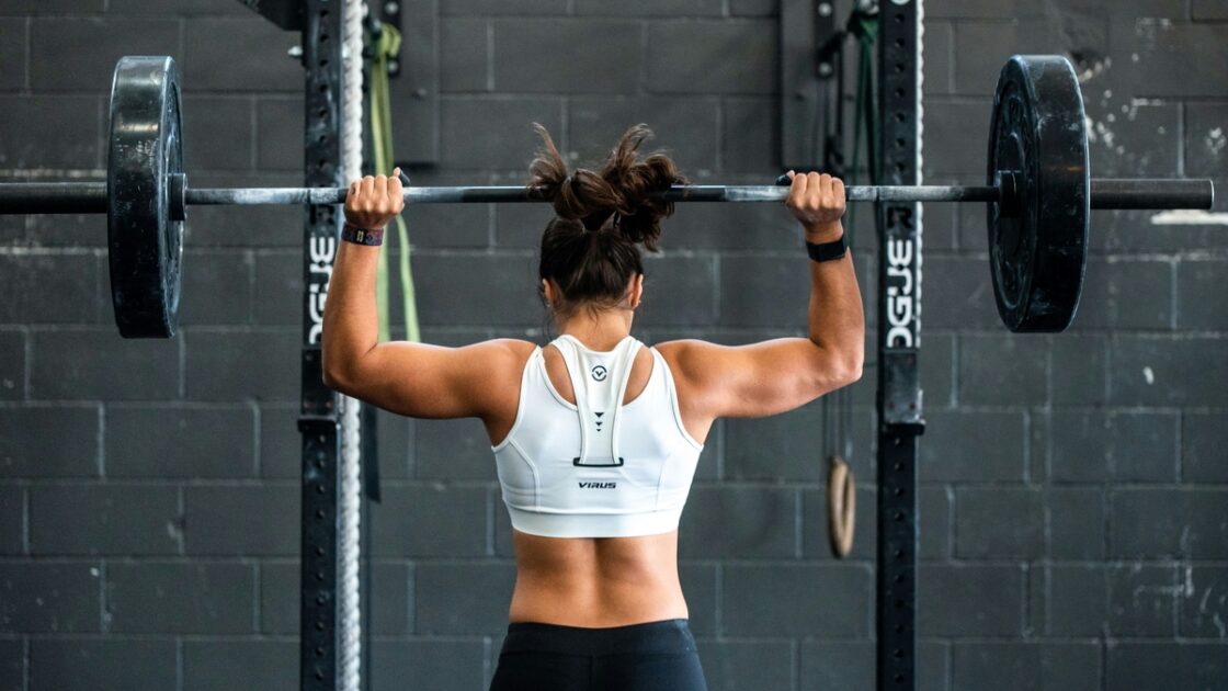Image of a young muscular woman with her back to the camera, lifting a barbell over her head at the gym.