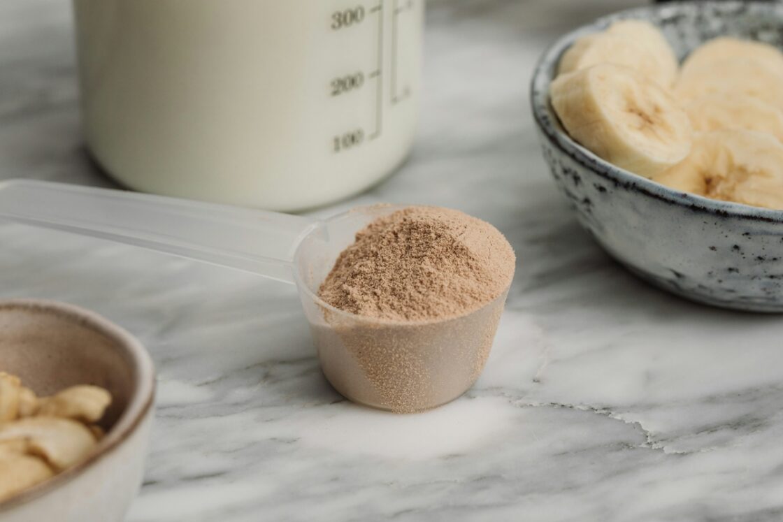 Image of a small plastic scooping spoon filled with an overflowing heap of chocolate protein powder, set on a marble countertop.