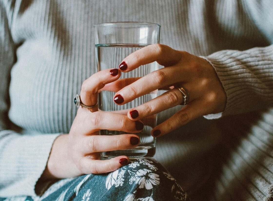 Image of a woman holding a glass of water with both hands, showing off a dark red manicure.