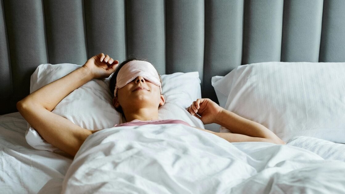 Image of a woman wearing an eye mask, laying down in bed and stretching her arms over her head.
