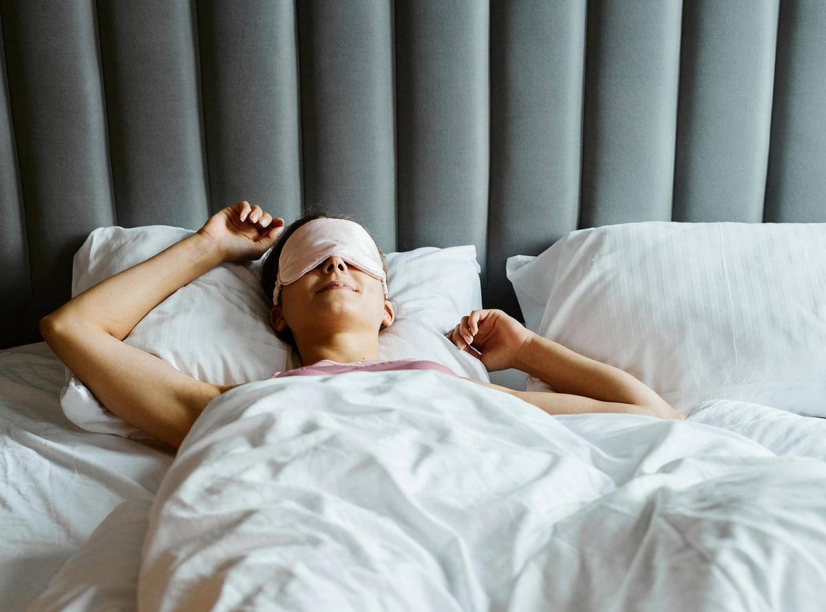 Image of a woman wearing an eye mask, laying down in bed and stretching her arms over her head.