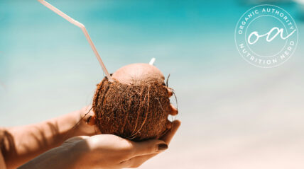 Image of a pair of woman's hands holding a brown coconut with a straw sticking out against an ocean backdrop.