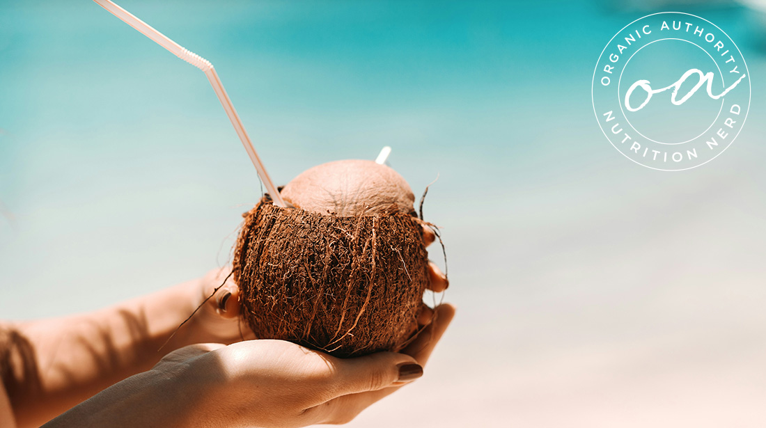 Image of a pair of woman's hands holding a brown coconut with a straw sticking out against an ocean backdrop.