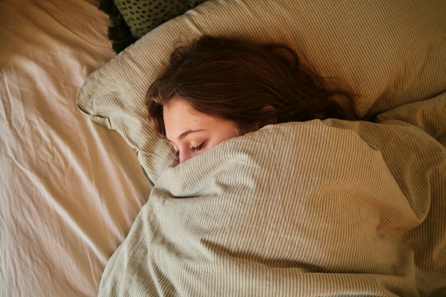Image of a young brunette woman sleeping under a beige duvet half covering her face.