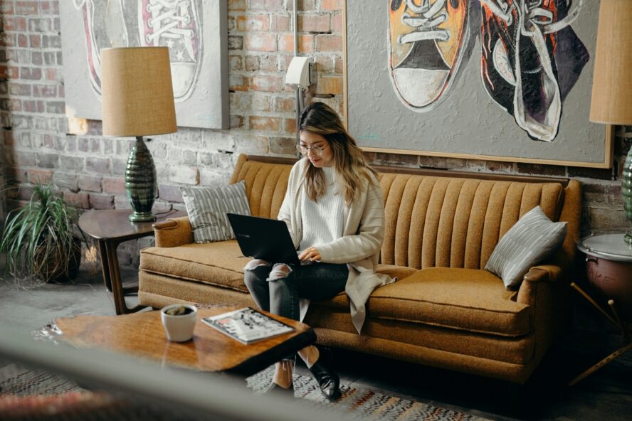 Image of a young woman sitting on a mustard yellow couch with a laptop perched on her lap.