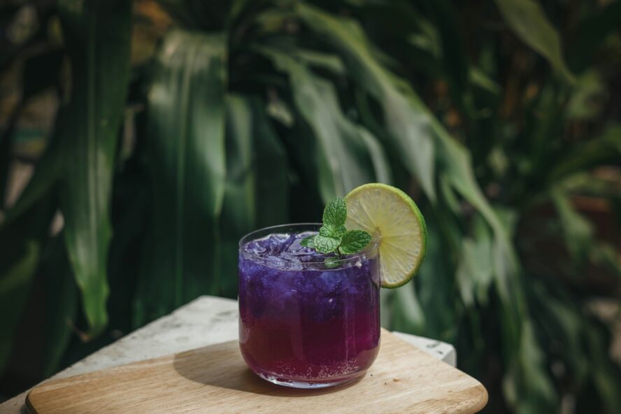 Image of an iced purple drink with a wedge of lime set on a white table against a leafy backdrop.