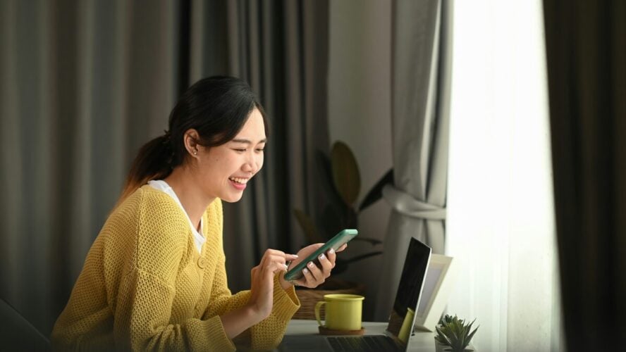 Image of a young woman sitting at a desk and smiling at her phone, with a laptop in front of her.
