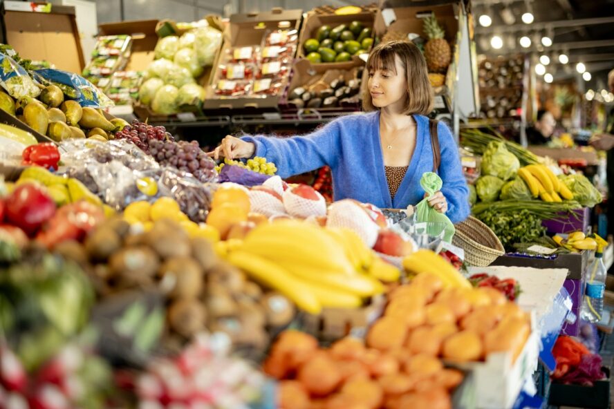 Image of a young woman with a bob and a blue sweater browsing in the fruit and vegetable section of the grocery store.