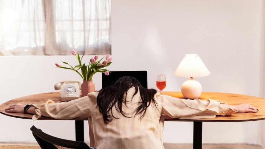 Image of a woman with her arms splayed on a desk in front of her laptop, with her head resting on the table.