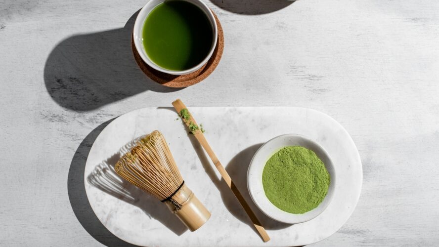 Image of a small bowl of matcha powder next to a whisk and small spoon, alongside a cup of matcha green tea.