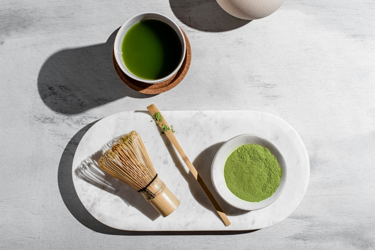 Image of a small bowl of matcha powder next to a whisk and small spoon, alongside a cup of matcha green tea.