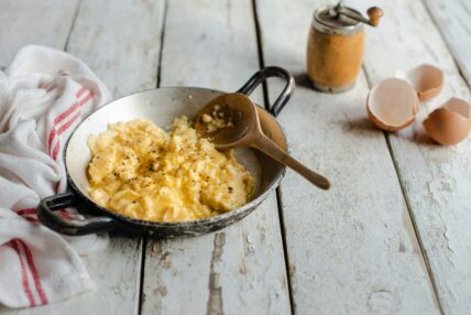 A small steel pan full of scrambled eggs with a wooden spoon, set on a rustic farmhouse table.