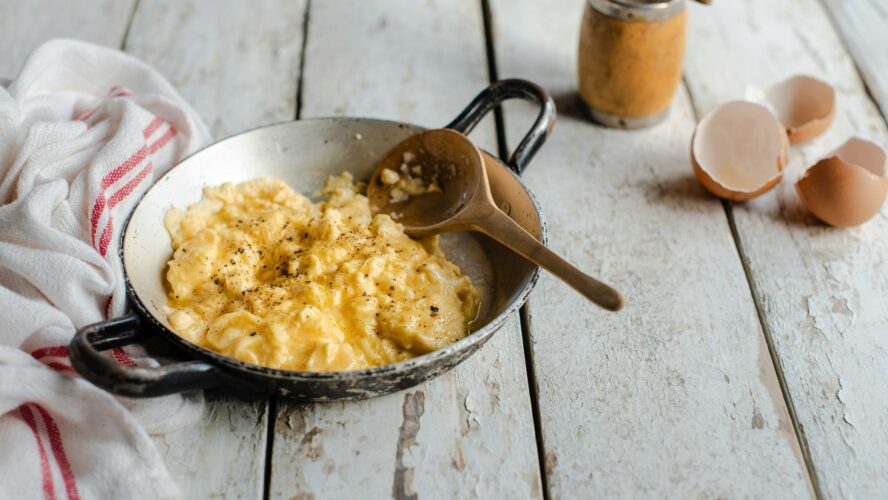 A small steel pan full of scrambled eggs with a wooden spoon, set on a rustic farmhouse table.