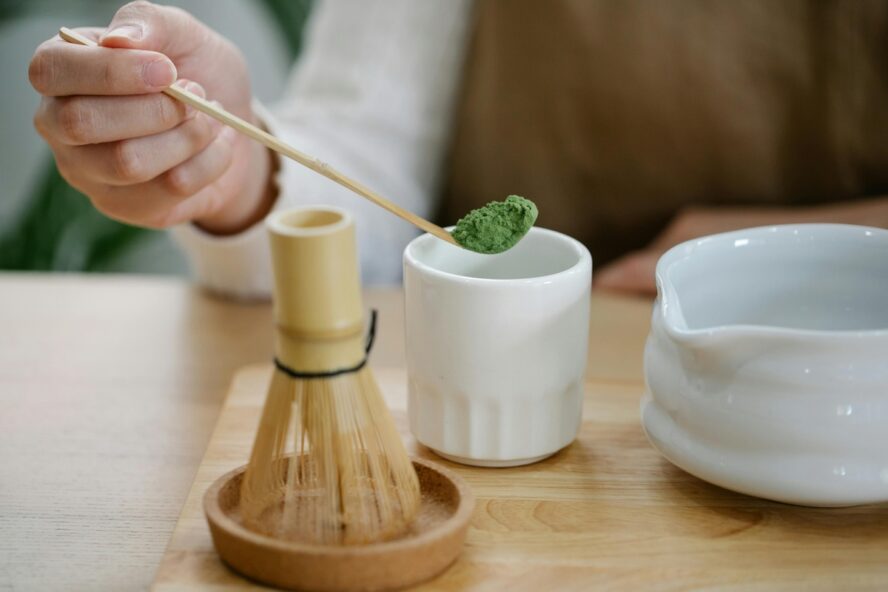 Image of a hand holding a small wooden spoon hovering over a cup, filled with matcha green tea powder next to a bowl and whisk.