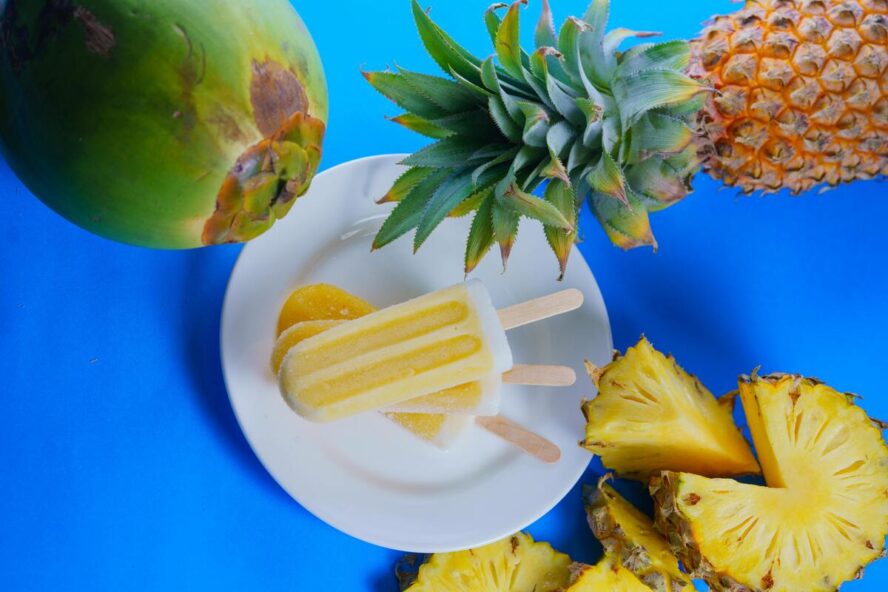 Image of two yellow popsicles on a plate on a blue background, next to coconut water and fresh pineapple.