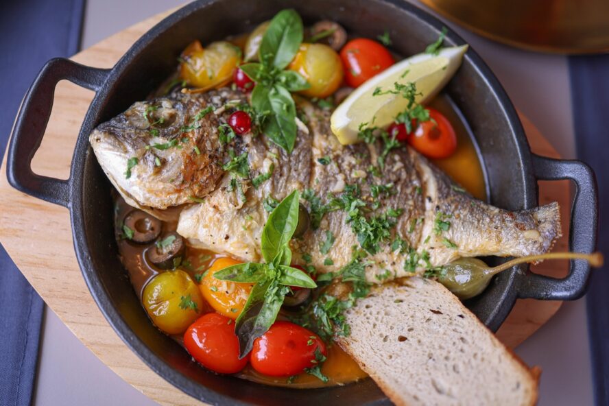 Image of a cast iron pan with a whole fish, cherry tomatoes, olives, lemon, and basil. 