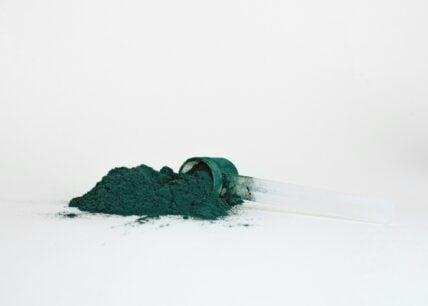 Image of a green-ish blue natural food dye powder spilling out of a small scoop onto a white surface.