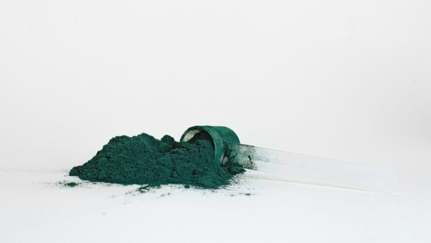 Image of a green-ish blue natural food dye powder spilling out of a small scoop onto a white surface.