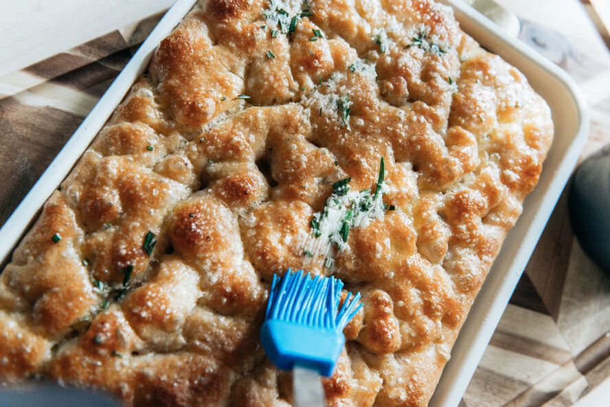 Image of herby garlic butter being spread onto a fresh pan of focaccia.
