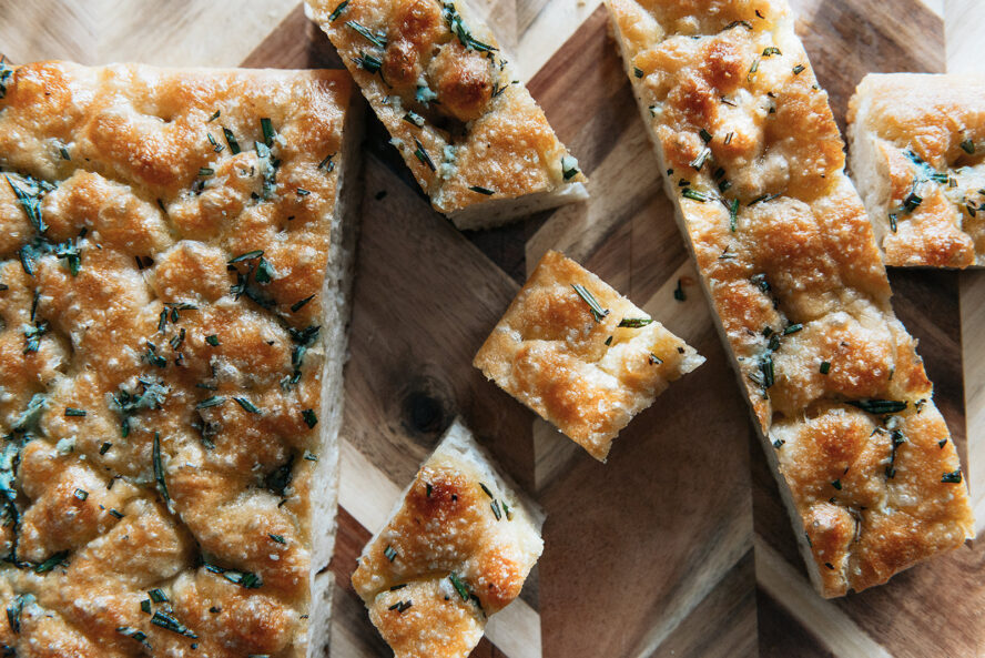 Image of a fresh pan of focaccia set on a wooden cutting board, cut into slices.