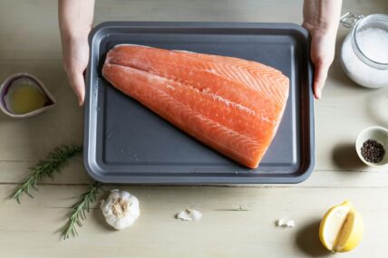 Image of a large filet of salmon on a baking tray held up over a counter with garlic, salt, and a wedge of lemon.