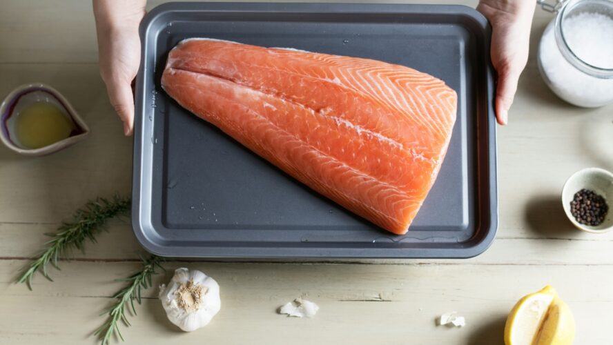 Image of a large filet of salmon on a baking tray held up over a counter with garlic, salt, and a wedge of lemon.