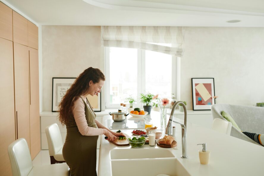 Image of a young pregnant woman standing at a counter in a bright kitchen, slicing some fruit.