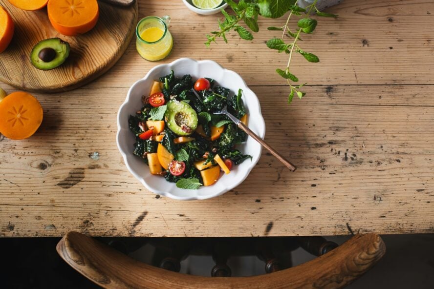 Image of a wooden table with a white ceramic bowl of cavolo nero salad with avocado.