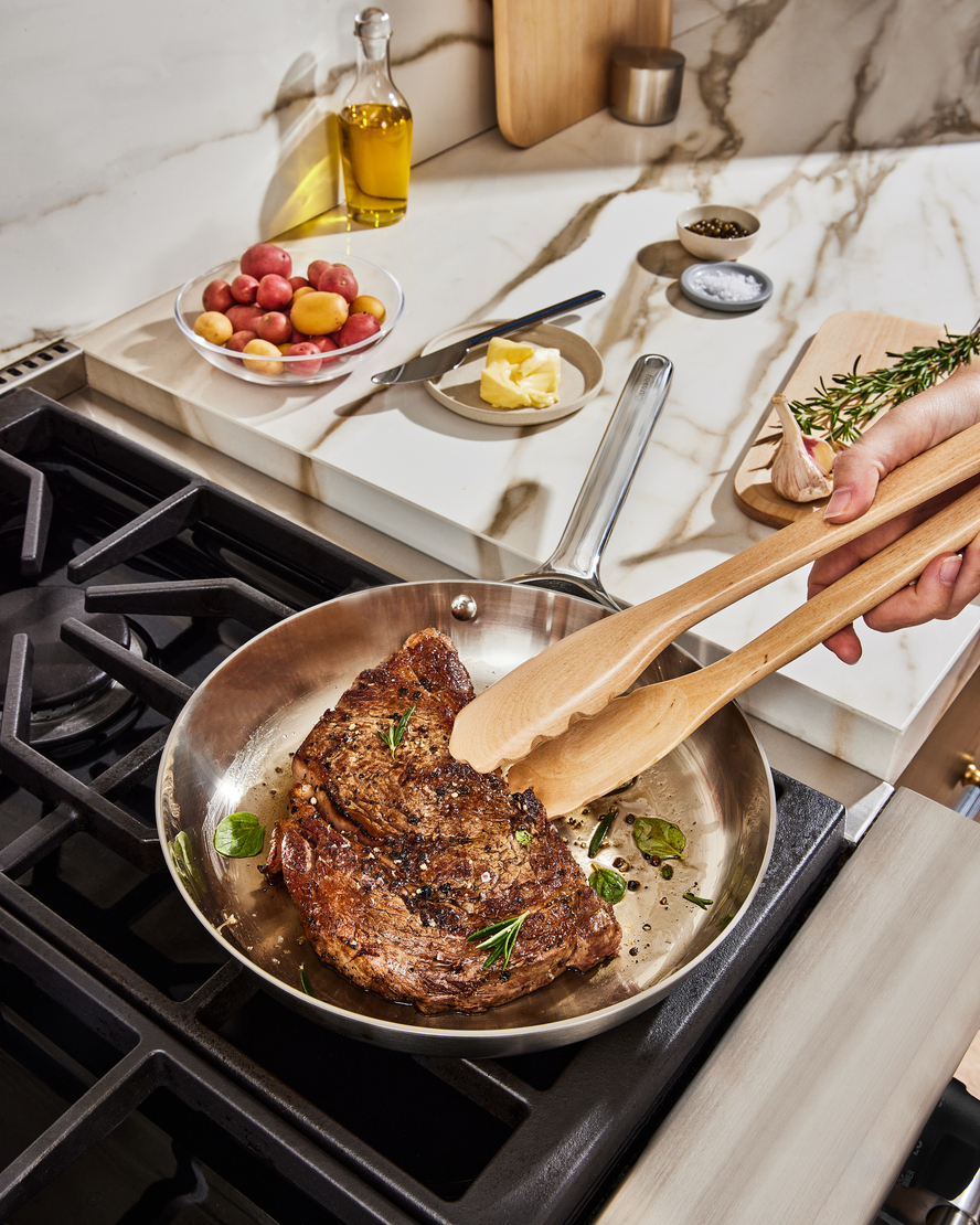 Image of a Caraway stainless steel fry pan with someone using a pair of tongs to sear a steak.