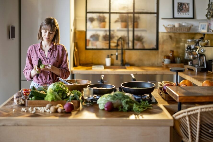 Image of a young woman standing at a kitchen island, cutting fruit and preparing lots of vegetables.