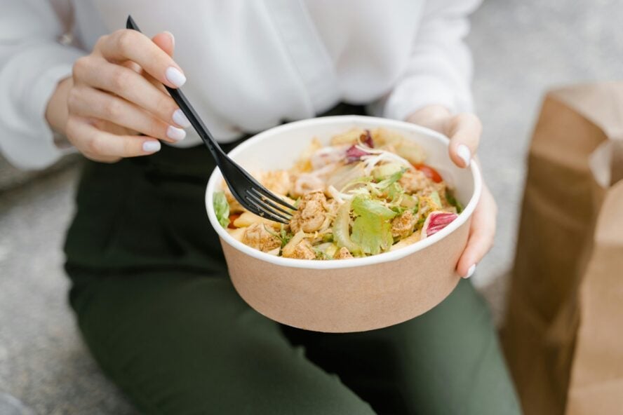 Image of a woman's hands holding a fork and takeout bowl filled with various grains and vegetables.
