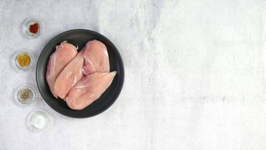 Image of several chicken breasts on a plate alongside several dishes of spices and herbs.
