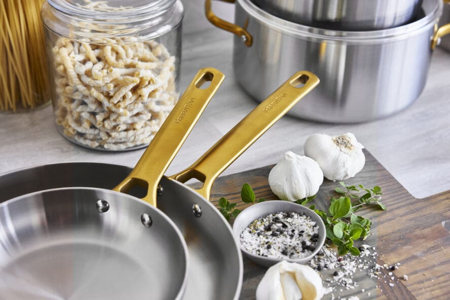 Image of two GreenPan Reserve Stainless Steel frypans nested in each other, with their iconic gold handles on display over a cutting board with garlic and truffle salt. 