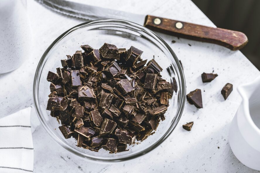 Image of a glass bowl full of chopped dark chocolate on a table with a white tablecloth next to a knife.