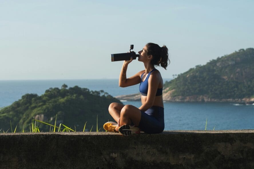 Image of a young woman in athletic wear sitting on a ledge, drinking from a stainless steel water bottle with a beautiful water view behind her.