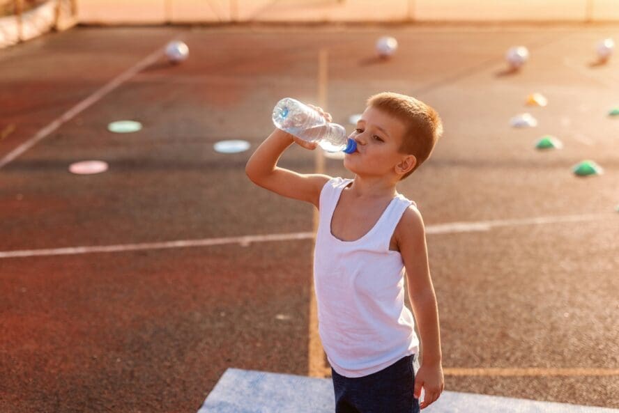 Image of a young boy standing on an outdoor basketball court, drinking from a plastic bottle of water.